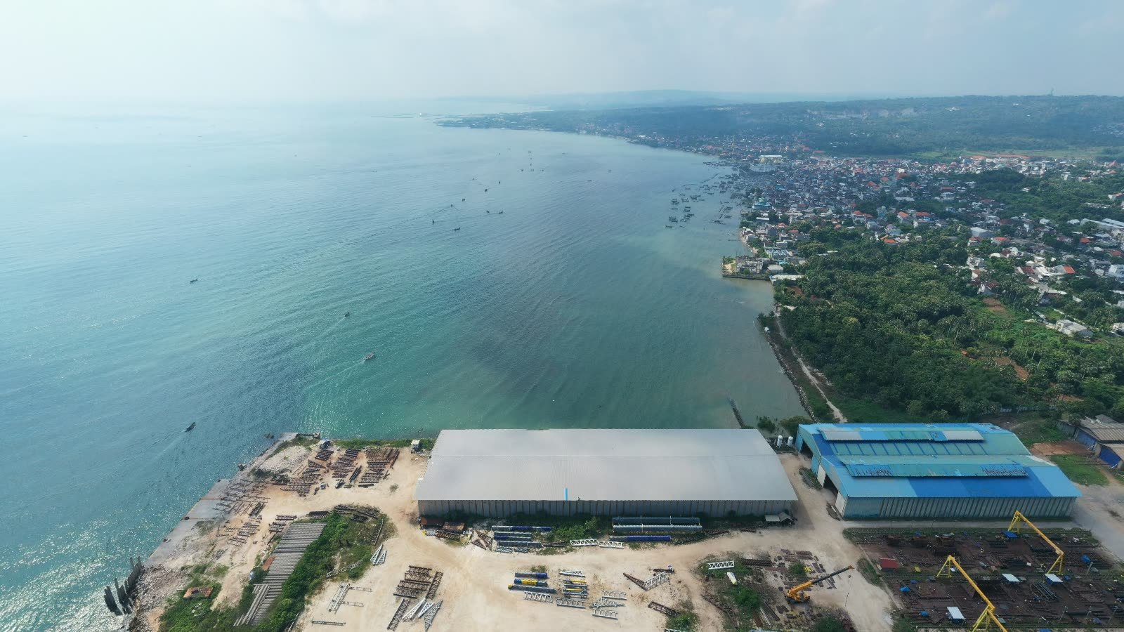 Lintech Seaside Facility dari udara: gudang panjang, bengkel beratap biru, dan crane portal kuning di tepi teluk, dengan kota Paciran di kejauhan.