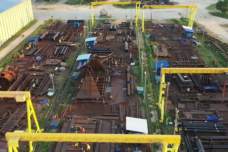 Bagian-bagian baja dirakit di bawah gantry crane kuning di halaman galangan, dilihat dari udara.
