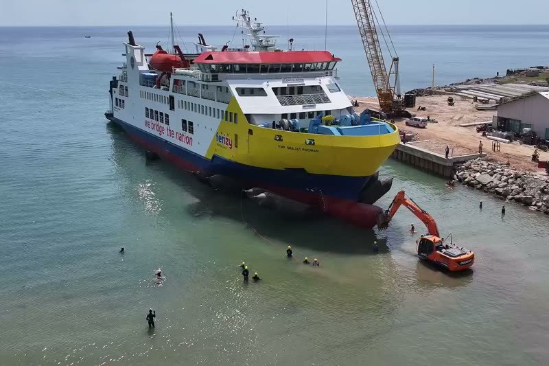 Kapal feri ditarik naik ke landasan galangan, dengan pekerja di air dangkal dan ekskavator di tepi.