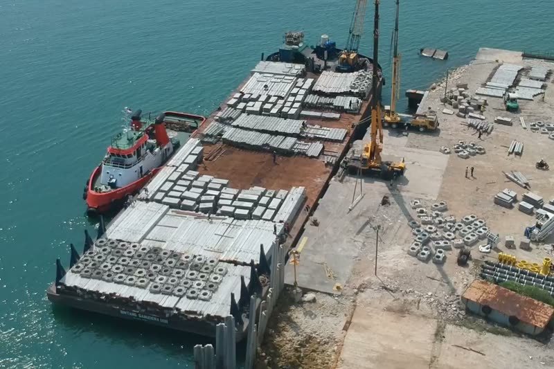 Kapal tunda di samping tongkang bermuatan blok beton di dermaga, dengan crane di tepinya.